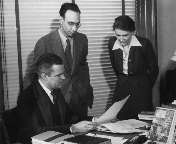 A man is sitting at a desk holding a document to show it to a man and a woman standing beside him. Caption reads: "Dr. Edward P. Alexander, Director of the State Historical Society of Wisconsin, Dr. Benton H. Wilcox, Librarian and Alice E. Smith, Director of Manuscripts Division. ca. 1942. Madison, Wisconsin."
