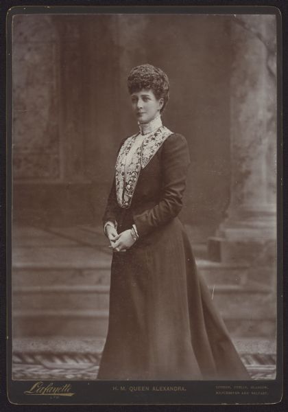 Three-quarter length standing carte-de-visite portrait in front of a painted backdrop of Alexandra of Denmark, Queen consort of the United Kingdom and the British Dominions, and Empress consort of India, 1901-1910. She was married to King-Emperor Edward VII.