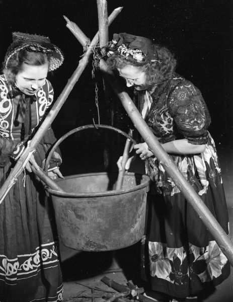 Two women in hats and dresses are stirring the contents of a cooking pot hanging on a tripod. Caption reads: "Stoughton, Wis. Pageant. Women in native costumes with old cooking pot."