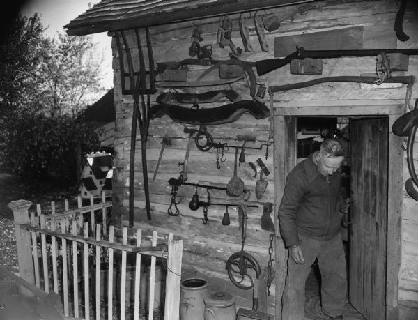 A man stands in the open doorway of a house. Many tools hang on the outside wall. There is a fence on the left, and a large birdhouse is at the corner of the log cabin. Caption reads: "McFarland (vicinity), Wis. Farm house filled with antiques."