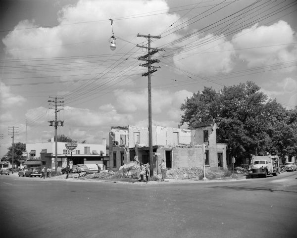 View across intersection towards several people standing outside a building that is partially demolished. A nearby sign advertises the Union House. Caption reads: "Madison, Wis. Razing/(?)old house on Milwaukee Street."