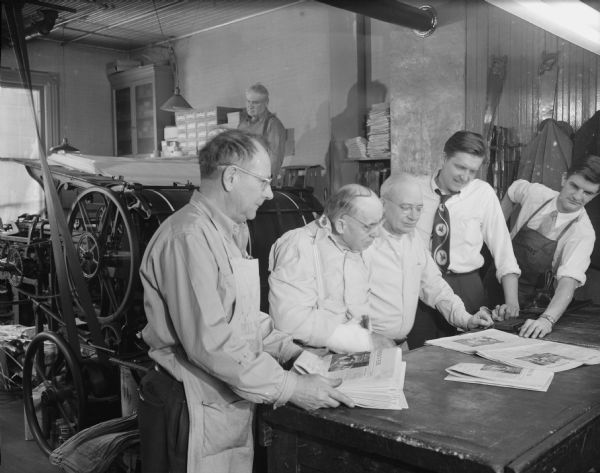 Five men are standing around a table, examining newspapers. A sixth man is running a press in the background. Caption reads: "Oregon, Wis. Oregon Observer, press room."