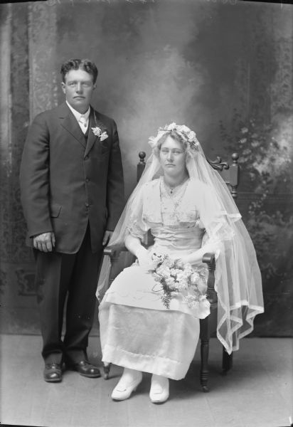 Studio portrait in front of a painted backdrop of a couple posed in a studio. The man is standing and the woman is sitting.