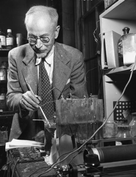 A man stands over a device, which he is inspecting. Caption reads: "Madison, Wis. 1949(?). Prof. Oliver P. Watts, retired distinguished authority on the corrosion of metals and electroplating, in his laboratory at the Chemical Engineering Department of the University of Wisconsin."