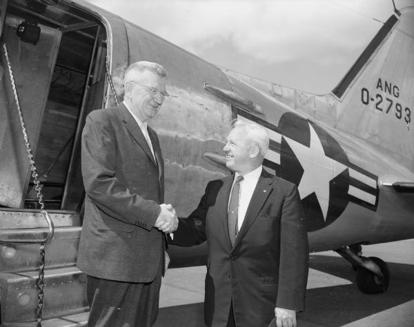 Two men smile and shake hands outside an airplane. Caption reads: "Madison, Wis. 1955-1956. Lieut.-Gen. Lewis Hershey (left) former head of the draft, and Brig.-Gen. [Ralph] Olson, Wisconsin Adjutant General."
