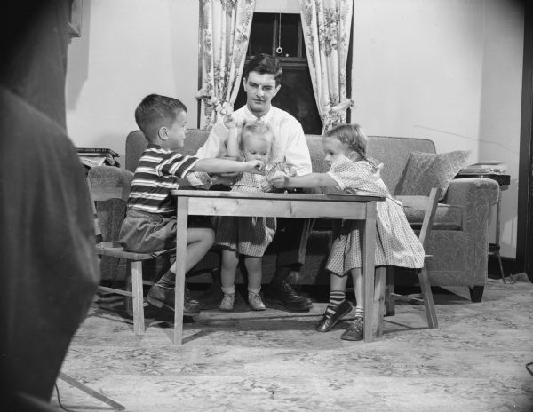 A man sits at a small table with his three children in a living room. Caption reads: "Madison, Wis. 1951-1952. Wallace Wicoff, chosen Man of the Year, with his children."