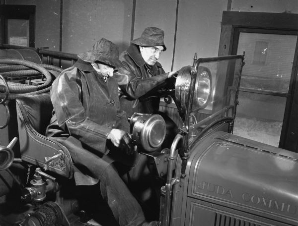 Two men sit in an old vehicle. Caption reads: "Juda, Wis. 1947-9. John Kryder, an old member of the local fire department."