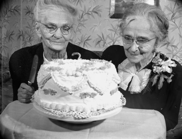 Two women smile and look at a cake; one is holding a knife. Caption reads: "Madison, Wis. 1947-9. Two ladies known as the "Bauhs twins" on their 83rd birthday."