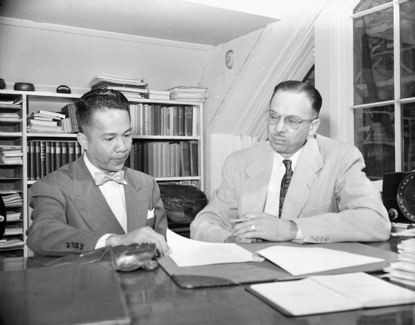 Two men sitting at a table, looking at a sheet of paper. Caption reads: "Madison, Wis. 1950-4. Harold Engel, assistant director of TV station WHA interviewing a visitor from Hawaii."