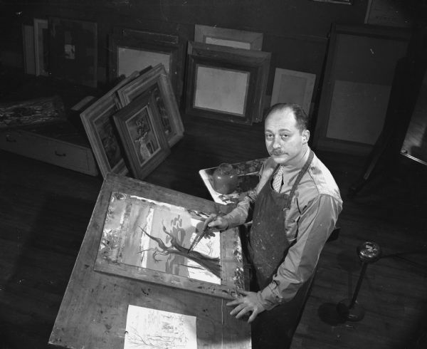 Elevated view of a man holding a paintbrush to a painting on a drafting desk. Several framed paintings are on the ground. Caption reads: "Madison, Wis. 1947-9. Aaron Bohrod, Artist in Residence at University of Wisconsin."
