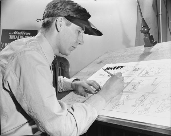 A man is working at a drafting desk on a multi-panel comic strip titled "Henry." Caption reads: "Madison, Wis. 1950-4. Carl Anderson, cartoon artist of popular comic strip character 'Henry.'" This caption, however, misidentifies the artist, who is actually Don Trachte. Trachte drew the Sunday version of "Henry" after the death of Carl Thomas Anderson. 