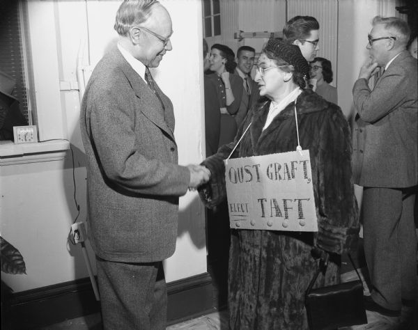 A man is shaking hands with a woman, who is wearing a sign that reads: "Oust Graft, Elect Taft." A group of people are talking the background. Caption reads: "Madison, Wis. 1952 (?) Visit of Sen. Robert Taft, candidate for Republican presidential nomination."