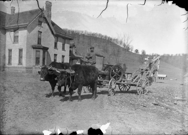 A man is posing standing on a cart behind two oxen, who are pulling a piece of farm equipment. A brick house is in the background, and behind the house is a hill. A sign painted on the side of the farm equipment reads: "Kissel, Wis" and "Hartf(ford)"?. Caption reads: "Lone Rock (?), vicinity, Wis. c. 1905-1910. Team of oxen pulling farm machinery."