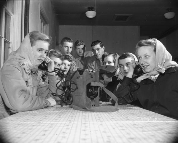 Six young women and three young men are sitting around a table. Two of the girls are using handsets from a telephone. Caption reads: "Madison, Wis. Civil Air Patrol training. Girls visiting(?)."