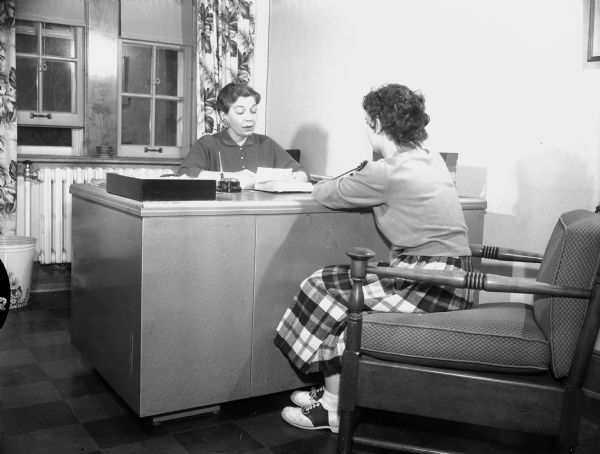 A teenage girl is sitting in a chair in the right foreground in front of a desk. A woman is sitting behind the desk facing her. Caption reads: "Oregon, Wis. 1956. State School for girls."