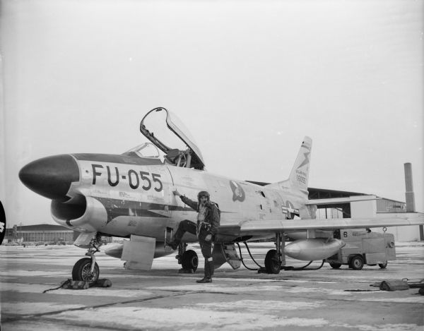 A pilot in flight suit poses next to an F-86D Sabre jet fighter. Caption reads: "Madison, Wis. Truax Airport. Pilot about to climb into a jet fighter."