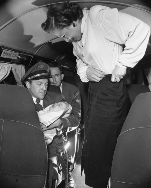 Two men and a woman are in an airplane. One man is sitting and holding a baby. Caption reads: "Madison, Wis. Wisconsin Central Airlines. Pilot or other personnel holding infant while in flight."