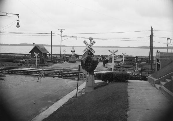 View of a railroad crossing near Lake Monona. Men are on the standing on the road in the background, and other men are working along the railroad tracks. Caption reads: "Madison, Wis. Law Park railroad crossing where man was killed."