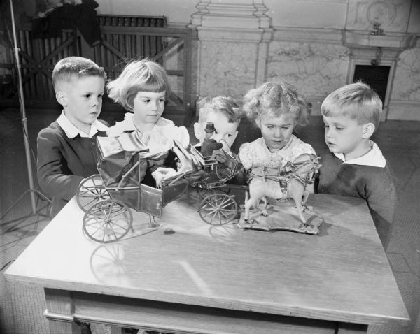 Five children are standing around a model on a table. Caption reads: "Madison, Wis. State Historical Society of Wis. Children looking at model of a horse and buggy."