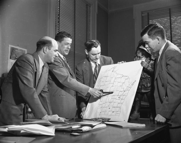 Four men are standing around a desk looking at a map of Wisconsin marked with the county borders. Clifford Lord is pointing at the map with a tobaco pipe. The map is supported from behind by a sculpture. Caption reads: "Madison, Wis. State Historical Society of Wis. Staff members John Jacques, Clifford Lord, Donald R. McNeil and Harry Hunter posing with a map.