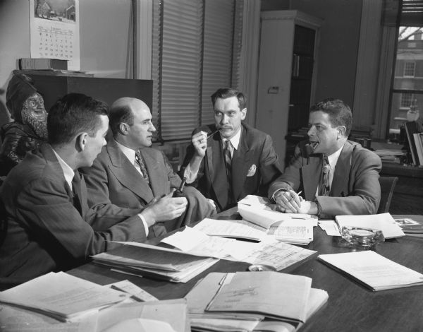 Four men are sitting around a table, three holding or smoking tobacco pipes. A sculpture is behind them on the left. Caption reads: "Staff conference. 1954. Left to right: Donald R. McNeil, John C. Jacques, Harry Hunter, and Clifford L. Lord."