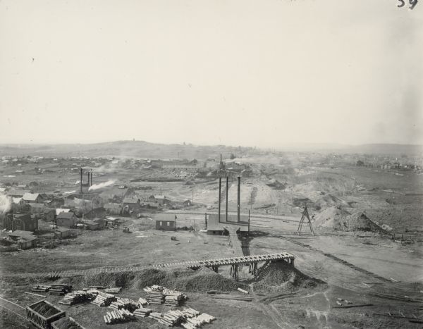 Elevated view of Hurley, WI and nearby industrial sites (Scott & Howe Lumber Co. Mill, Ashland Mine). Inventory reads: "Looking west and a little south over the Ashland Mine & Scott + Howe and you see the Hurley water tank on Germania Hill and the Iron Co. [County] Courthouse tower in the background." Caption reads: "Abt 1907 - The steeple on the Hurley cathedral is incomplete."