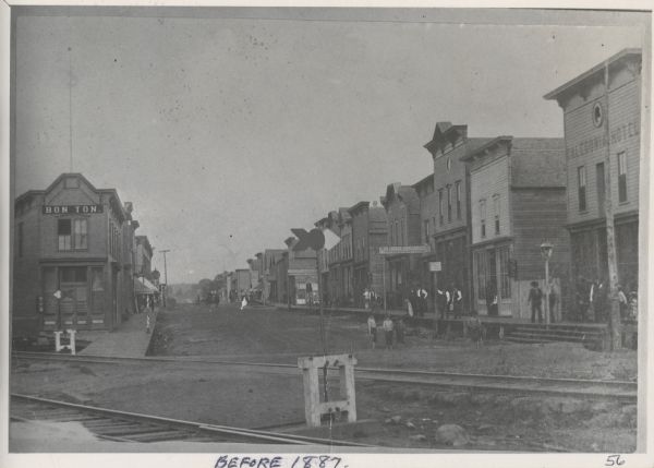 A view of  the businesses present on Silver Street, Hurley's main street, less than three years after the city's settlement. City residents and a horse-drawn carriage are in the street. Caption written by Gib Endrizzi: "Hurley, looking east from the depot (corner of 5th Ave. and Silver St.), 1887. Within one year from the start of construction, Hurley's Main Street showed remarkable progress. Commenting on the development of Hurley, the Gogebic Iron Tribute noted, 'Each day, a new building goes up, and the woods resound with the clash of falling trees and the music of a saw and hammer.' By 1887, Silver Street was packed with storefronts along its five blocks with many more businesses on side streets. On June 7, 1887, a fire that started in the Gogebic Meat and Provision Company destroyed all of Silver between 2nd and 3rd Avenues. A second, more disastrous fire started in the Alcatraz Theater (the tallest building at right) on July 9. Most of Silver Street to the east and many of the buildings seen in this photo, including the Bon Ton and Caledonia Hotel, were consumed in the fire."