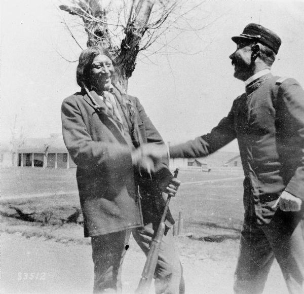 Two men smiling and shake hands outdoors. The man on the right is wearing a military uniform, and the man on the left is holding a rifle. There is a building in the background. Caption reads: "[Arapaho chief] Sharp Nose and Capt. Wm. C. Brown. 1st Cavalry at Fort Washakie, Wyo., 1899."