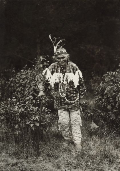 Portrait of a man standing in a clearing. He is wearing a headband with two feathers and necklaces, and is leaning on a cane. He is identified as Chief Pussafiss, an Ojibwe chief who lived in Solon Springs and died in January 1913.