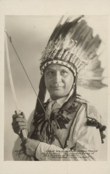 Quarter-length portrait of a man wearing a feathered headdress, claw necklace, and vest. He is holding a bow. Caption reads: "Chief Whitefeather — Indian Mound Reservation — The Summer Camp of Boy Scouts of America Milwaukee County Council." Caption on back reads: "John Gosling (White Feather). Chippewa Indian. Director, Woodcraft, Boy Scout Camp. Silver Lake, Oconomowoc. July 1927."
