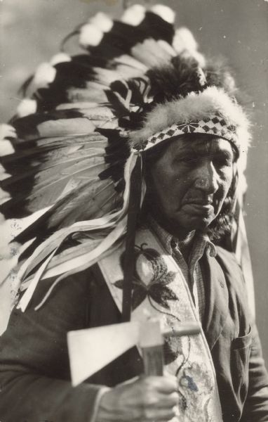 Waist-up portrait of a man wearing a feathered headdress and holding a tomahawk. Caption on back reads: "Chief Greenhill, 'Jim Goose,' Chippewa Tribe, Cass Lake Band. Died 2/5/38."