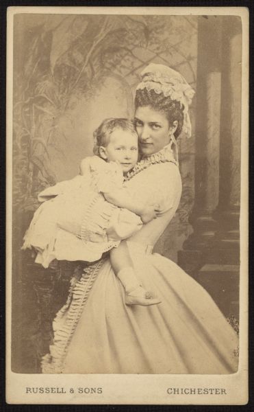 Carte-de-visite portrait in front of a painted backdrop of Alexandra, Princess of Wales, later Queen of the United Kingdom, posing with a toddler in her arms. This is probably her first-born son, Albert Victor.