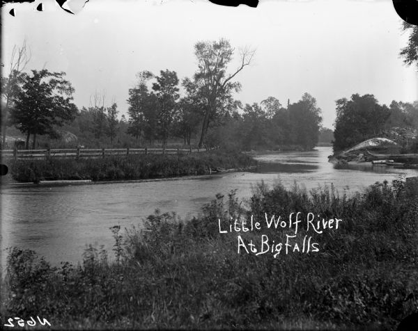 View from shoreline across a river. A wooden fence is along the far bank. Caption reads: "Little Wolf River At Big Falls."