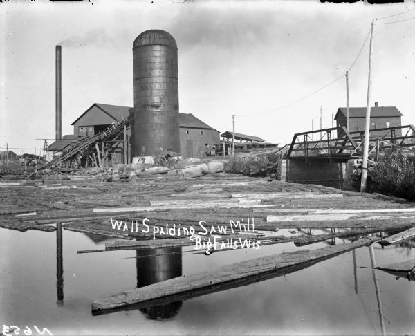 View of a saw mill and silo. Several logs are floating on the river in the foreground. A bridge is on the right. Another building is in the distance. Caption reads: "Wall Spalding Saw Mill, Big Falls." 