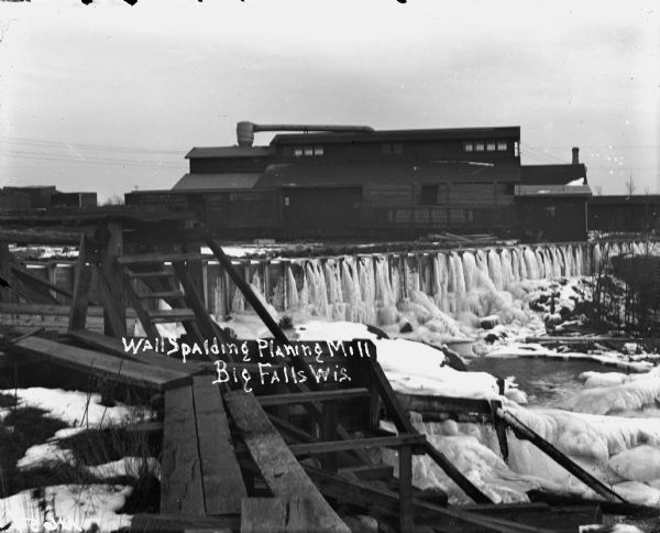 View across water towards a large building above a waterfall. Ice is coating the sides of the river. A bridge is in the foreground on the left. Railroad cars are along the side of the building near the waterfall. Caption reads: "Wall Spalding Planing Mill, Big Falls Wis."