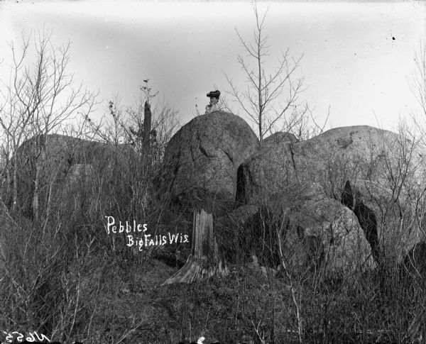 View of several rounded rock formations amid trees. A woman wearing a hat is posing behind one large rock in the center. Caption reads: "'Pebbles' Big Falls, Wis."