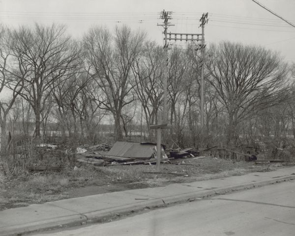 View from street towards a building that has collapsed. There is a sidewalk in the foreground, and trees surround the building. A body of water is in the background.