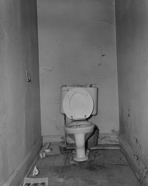 View of a toilet in a dirty stall. The plaster on the walls appears damaged, and the broken toilet tank lid is laid next to it. 