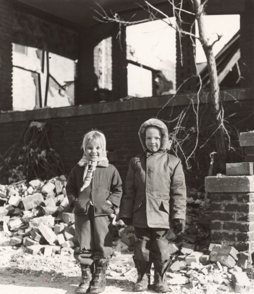 Two children in winter coats and boots posing outside a building. Bricks are scattered on the ground behind them, and the building has cracked windows.