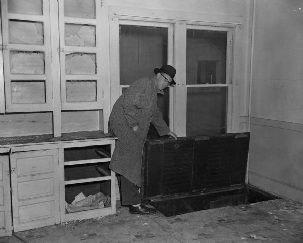 A man in a hat and coat posing opening a cellar door inside a building. Bare shelves and cupboards are beside him.