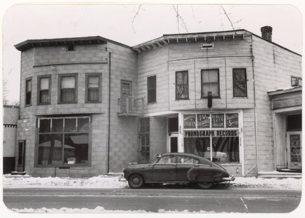 View of a building exterior from across the street. Signs in the shop window read: "Wholesale prices," "TV exchange," "As-built sets $40 & up," "Radios $5 & up," and "Phonograph records." A car is parked outside the building. This location is identified by the Madison Redevelopment Authority as "Project Parcel no. 16-10."
