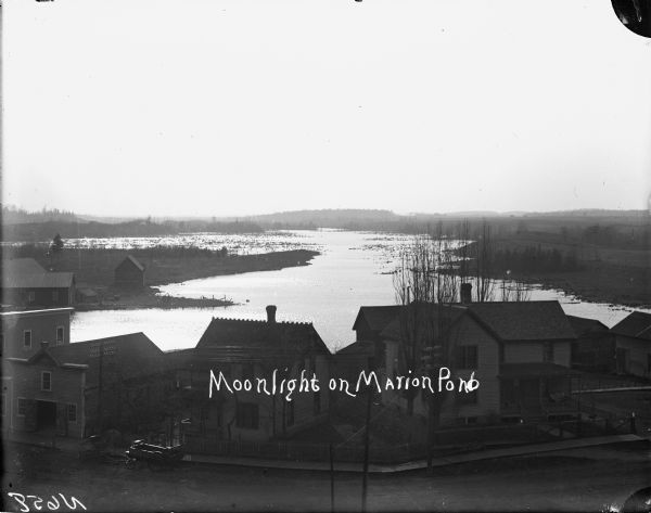 Elevated view over the tops of buildings to the water beyond. A wooden sidewalk and fence runs along the street in the foreground. Caption on negative reads: "Moonlight on Marion Pond."