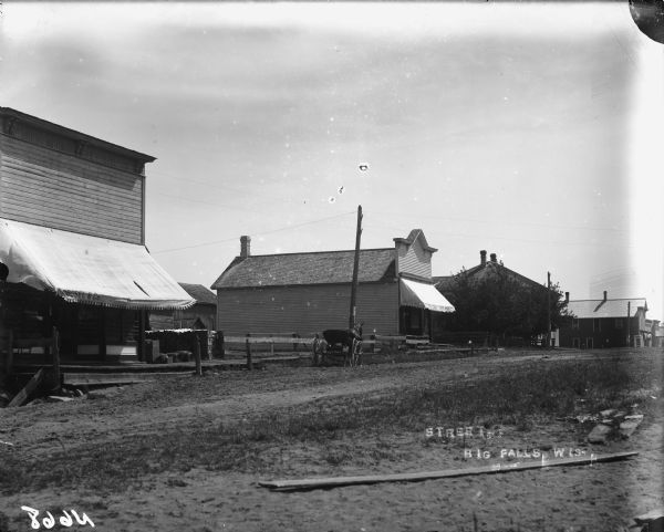 View across unpaved street toward several buildings, and a horse-drawn buggy near a fence. The building on the left has an awning with the sign "[?]obosel and Co." Caption on envelope reads: "Street in Big Falls, Wis. 1890-1910?" Further down the street in the center is a building with an awning that reads: "Cigars" on the side, and on the front: "? Toomey Choice Wines."