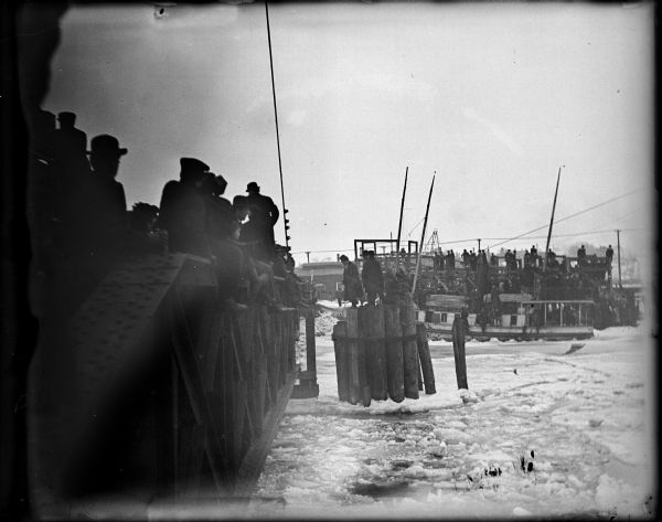 Caption reads: "Manitowoc, Wis.? 1906. Passengers disembarking ship in winter." A group of people are sitting and standing at the side of a bridge in the left foreground, and a building is on the snowy shoreline just beyond the bridge. Another group of men are standing on pilings in the icy water near the bridge. In the background people are on a ship and behind the ship people are standing on a pier or bridge.
