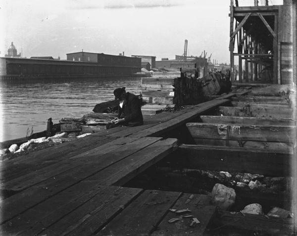 View towards a man sitting on a dock. A pipe is laying on the dock beside him. On the opposite shoreline are long industrial buildings. Caption on envelope reads: "Manitowoc? Wis. ca. 1904-1908. Old man sitting on port dock."
