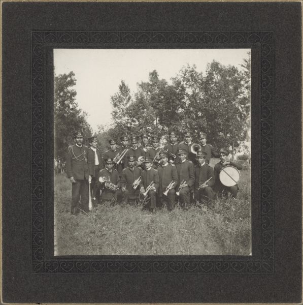 Group portrait of a band posing in a field with trees in the background. All the men are wearing uniforms and holding their instruments.