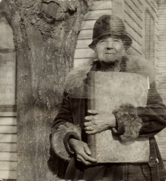View of a woman holding a large bound volume and standing next to a tree. Caption reads: "An Indian woman of over 70 [Lura Fowler Kindness] who lives by herself in a little house on the main highway in Brothertown. She has a ms. book in her possession — a record of the Brothertown Indians as written by their superintendents since 1795. She will not part with it or send it for photo-stating. Willing to have some one come to her home to copy. Apr. 10, 1934 Mr. Schafer told her that if he should come to that part of the State during the summer he would come to see her. Letters in office file:
Coe Hayne - Mar. 27, 1934
Mrs. Kindness - Mar. 30, [1934]."