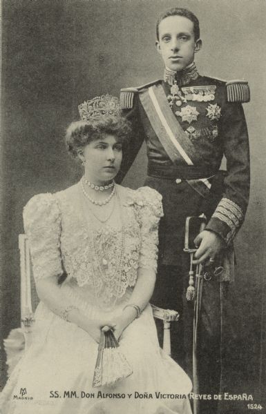 Portrait of King Alfonso XIII and Queen Victoria of Spain. He is standing and wearing a military dress uniform with sword, and she is sitting and wearing a gown and tiara and holding a fan. Caption reads: "SS. MM. Don Alfonso y Doña Victoria Reyes de España."
