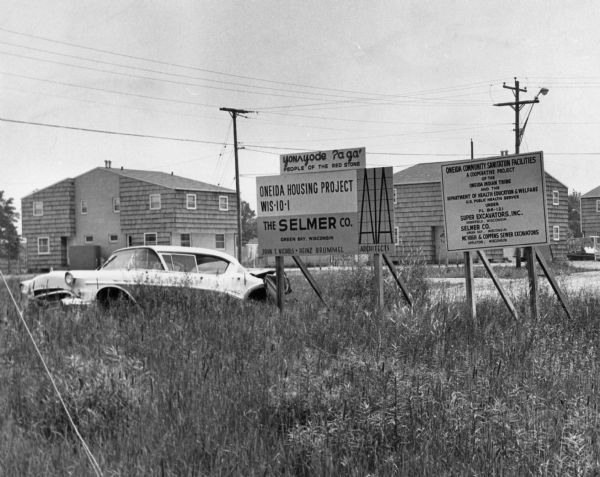 View of signs in a field near a damaged car. There are buildings in the background. Signs read: "yon&#652;yode &#704;agá People of the Red Stone," "Oneida Housing Project WIS-10-1," "The Selmer Co. Green Bay, Wisconsin John T. Nichols - Heinz Brummel Architects," "Oneida Community Sanitation Facilities A Cooperative Project of the Oneida Indian Tribe and the Department of Health Education & Welfare U.S. Public Health Service under PL 86-121 Super Excavators, Inc. Brookfield, Wisconsin Selmer Co. Green Bay, Wisconsin, McHugh & Coppens Sewer Excavators Appleton, Wisconsin." 