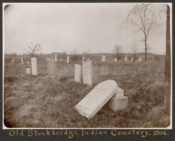 View of a cemetery with several tombstones, one of which appears broken and resting against its base. A building is in the background. Caption reads: 
"The Old Stockbridge Indian cemetery is on Lot 89, is no longer used, is surrounded by a barb wire fence with no entrance gate. It is in a very neglected condition, with many tomb stones fallen down or leaning over, some broken, others cracked and all overgrown with moss, making the inscriptions often illegible. Was made use of as a cow pasture by the present owner for some time, but owing to remonstrance by the Indians, is no longer put to that use. 
The farm house in the distance is that of a Stockbridge Indian. The tombstone in the foreground is that of Austine Quinney, the old chirf [sp], who signed the Articles of Agreement at Vernon, New York, in 1823 (see <u>Wis. Hist. Colls.</u>, XV p. 8).
Some other names inscribed upon the 150 to  200 graves are:
Percillla Quinney
Washington Quinney
Darius Charles Quinney
Amelia Quinney
Olive P. Quinney
Chloa M. Crosley
Joshua Wilson
Amzi C. Wilson
Diademia Miller
Delilah Miller
Thomas S. Shenandoah
Tietta Moore
Lewis Boman died July 14, 1858
Eunice P. Abrams died 1846
Lucy Pye
Irenaia Pye
Charitty A. Palmer"
